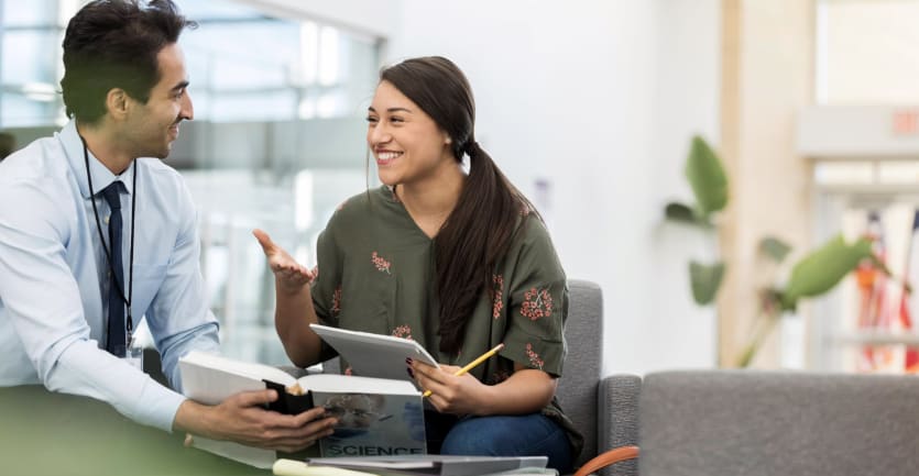 A female college student happily chats with her professor mentor, who cheerily refers to a textbook that reads SCIENCE on the cover.