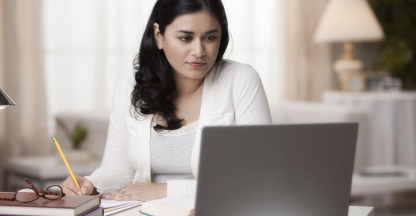 A woman in a white cardigan studies intently from her laptop computer, pencil in hand, as she sits at her dining room table.