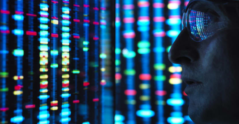 A scientist gazes at a screen displaying color-coded genetic patterns, the images reflecting off his glasses.