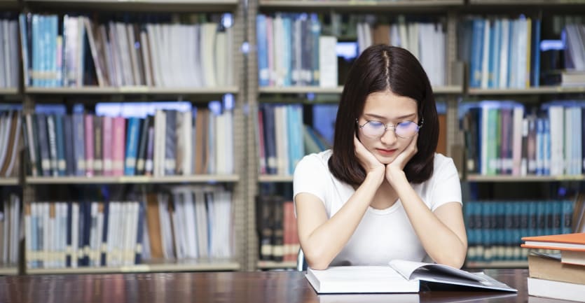 A woman withround glasses and a white shirt sits at a conference table in a library, staring intently at an open book while proppingup her chinwith her hands.