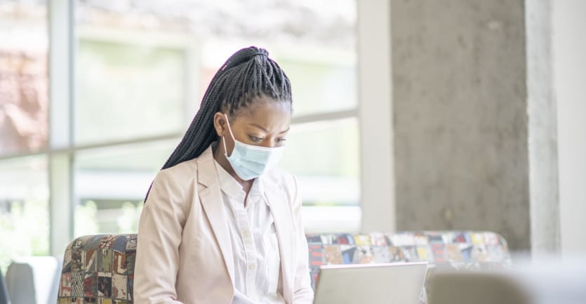 A woman in a white button-down, collared shirt, a cream-colored blazer, and a light blue face mask sits alone on a couch in an academic building, typing on her laptop computer.