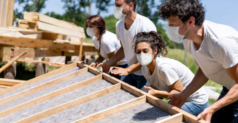 Four young adult volunteers in matching blue jeans, white t-shirts, and face masks work together to hoist up the wall of a home the group is building.