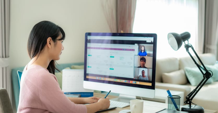 A woman in a ribbed pink sweater takes handwritten notes as she participates in a video conference on her desktop computer.