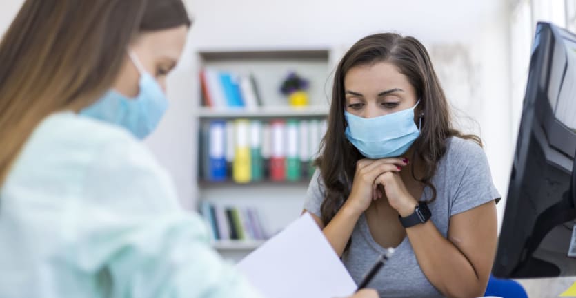Two women wearing face masks sit at a conference table and confer over miscellaneous printed materials.