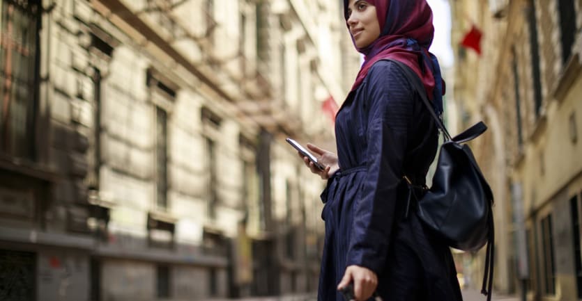 A young woman donning a pink and purple headscarf and a black backpack stands in the middle of a city, holding a cell phone in one hand and clutching a suitcase with the other.