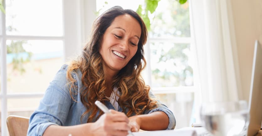 A woman in a denim shirt with rolled-up sleeves, sitting at her kitchen table, smiles as she takes hand-written notes next to her laptop computer.