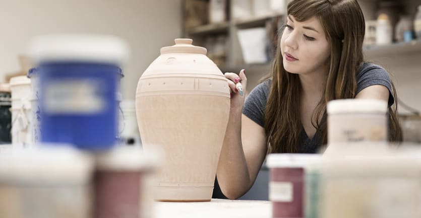 A young woman in a dark-gray t-shirt gingerly handles a clay urn in an artist's studio.