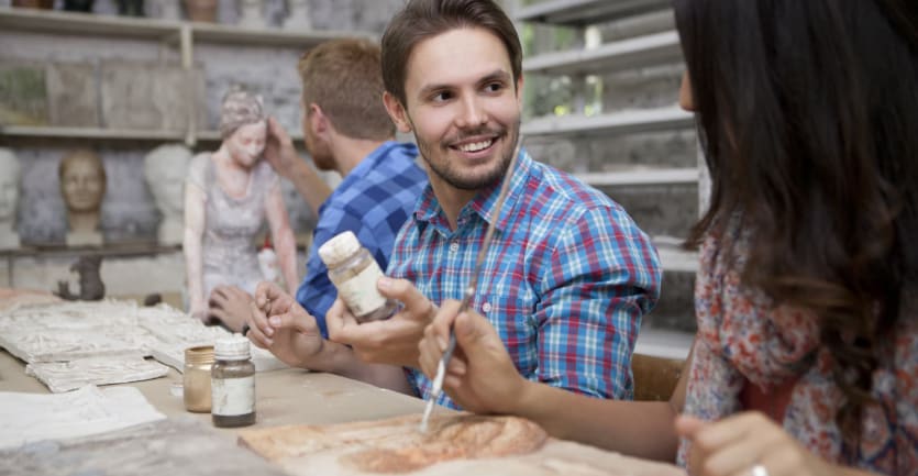 A man in a brightly colored plaid shirt smiles as he chats with a fellow student while seated at a table in a pottery studio.