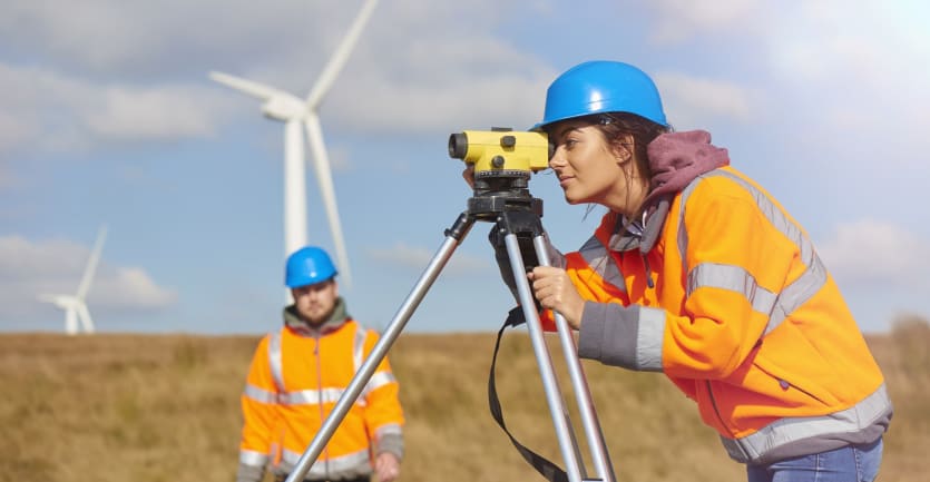 A woman in an orange jacket and blue hardhat peers through the scope of a level as her coworker walks toward her through an open field, wind turbines churning in the background.