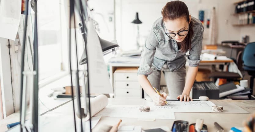 A woman wearing glasses, in gray denim jeans and a shirt with rolled up sleeves, leans over her desk to look at architectural plans.