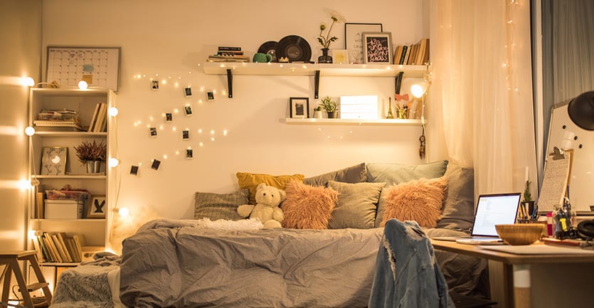 One side of a dorm room is presented in tasteful neutral neutral colors: a simple modern white desk, a bed covered in pillows and a duvet cover, string lights dangling from shelves.