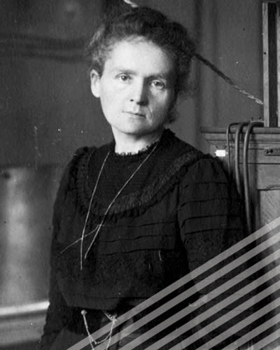 A black-and-white photograph shows Marie Curie in a black dress standing alone in her studio, eyes a little tired, as she looks directly into the camera.
