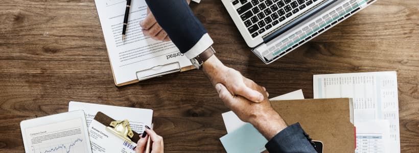 Best Online Master's in Business Administration (MBA) Programs. Above angle image of business meeting with associates shaking hands over charts, documents, and a laptop.