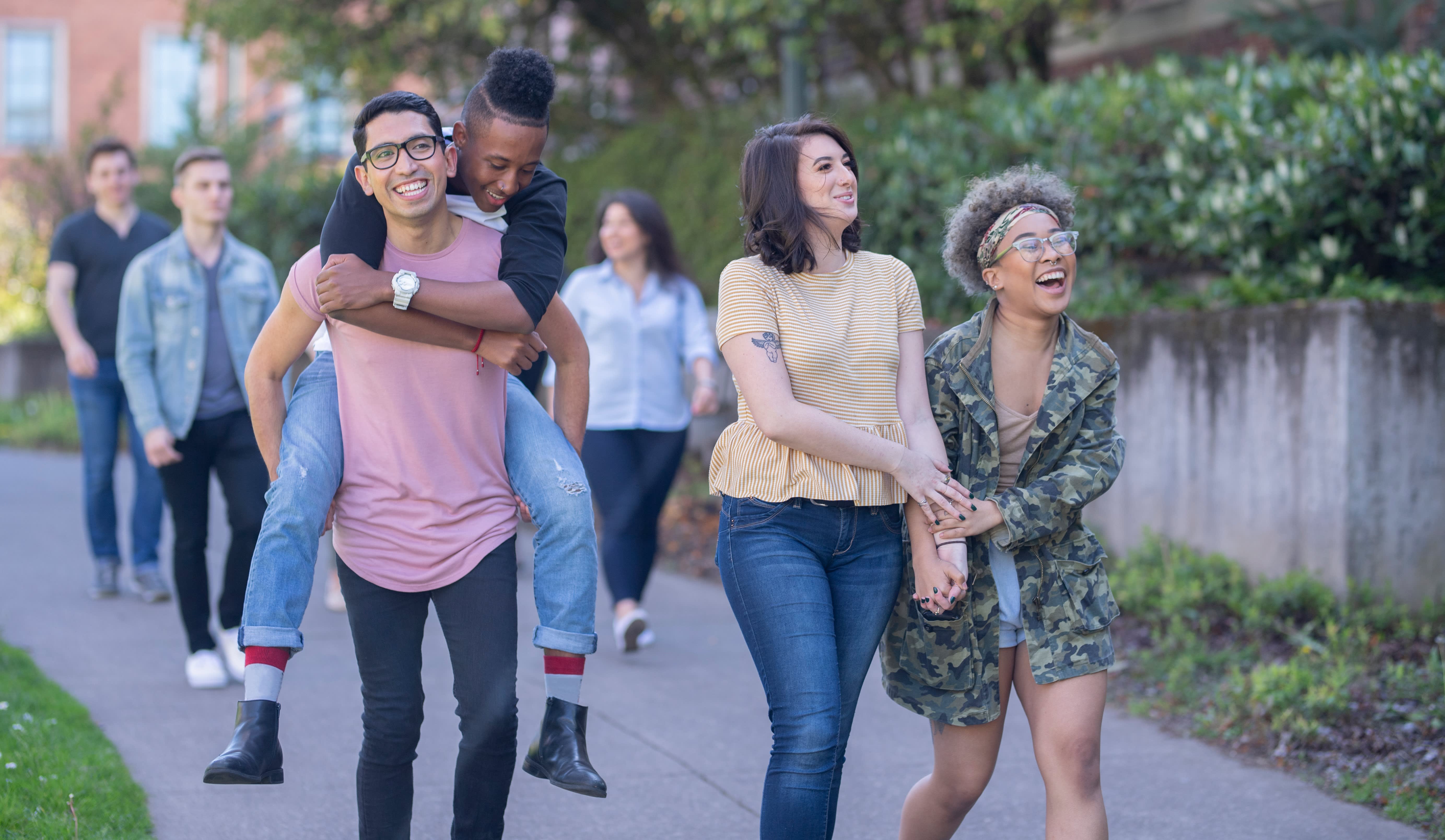 Two couples walk through a college campus, with one doing a piggy back ride