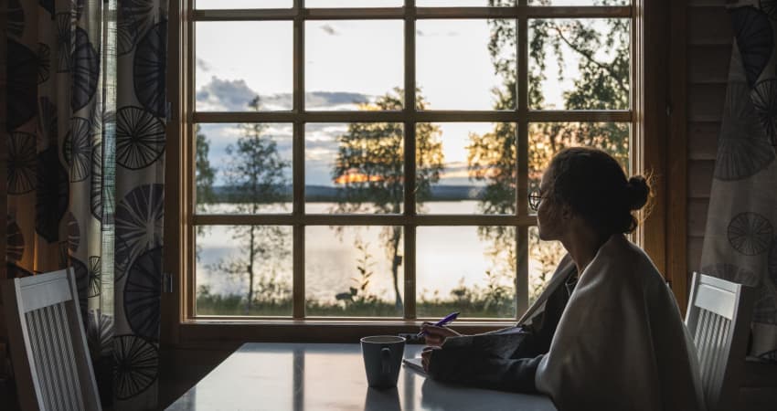 A person sitting at a table with a notebook stares out the window at a lake