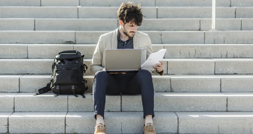 A student sits on cement stairs with his backpack and laptop, looking at a printed resume