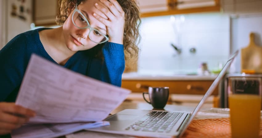 A student sits at a desk next to a computer, staring with concern at a bill