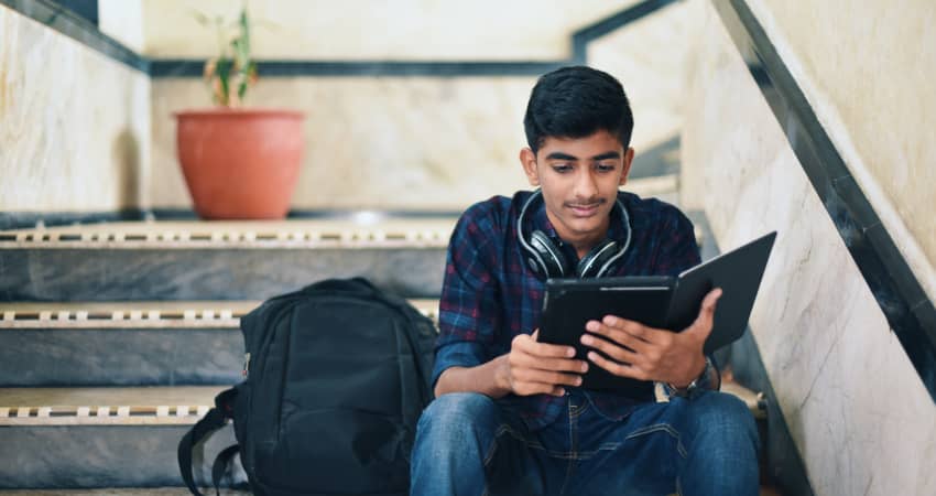 A student sits on marble stairs next to a backpack, looking at his tablet computer