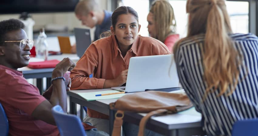 Three students sit around a desk in a classroom, discussing their work