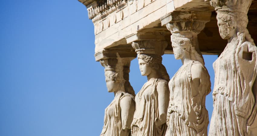 Four crumbling marble statues of women in robes decorate the columns of a Roman ruin, white against a blue sky