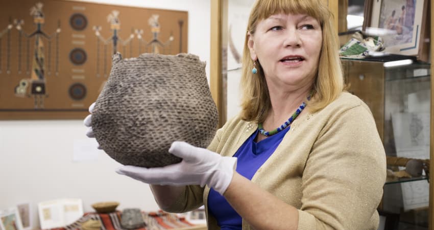 An archivist wearing white gloves holds up an old, woven basket in a museum collections room