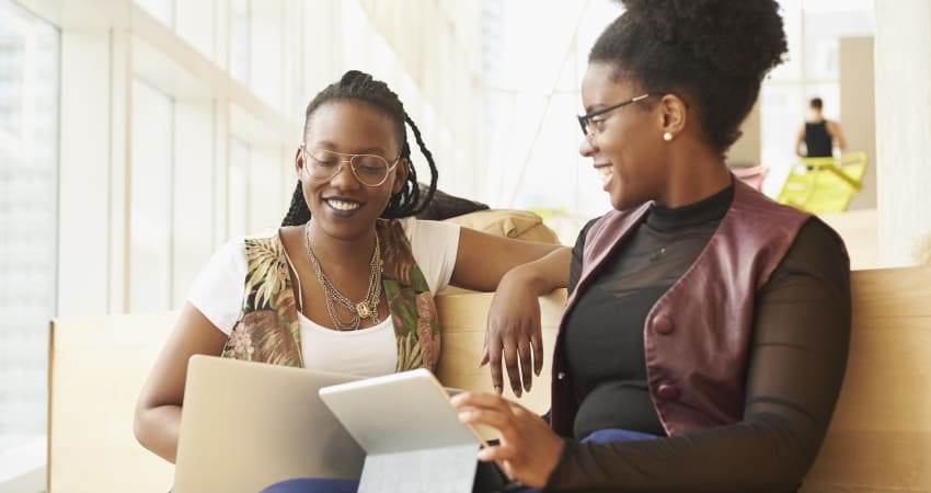 Two women sit on a bench, consulting over their laptops