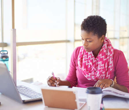 A young, black female college student is studying for her HESI exam. She's wearing a bright pink shirt and scarf. She is taking notes from her textbooks, and her laptop is open. Image credit: AJ_Watt / E+ / Getty Images