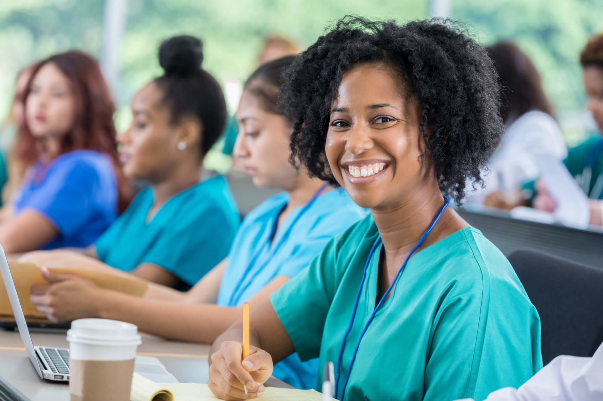 A smiling mid-adult African American nursing student is sitting in class with her colleagues. She has short, curly black hair, and is wearing teal green scrubs. Her laptop and notepad are open so she is ready to take notes during class. She is looking directly at the camera.