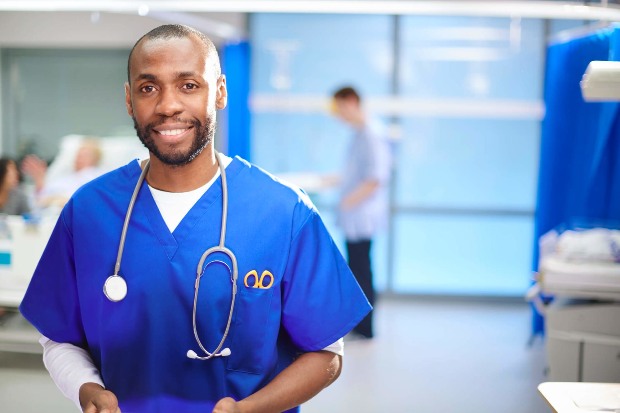 A mid-adult African-American nurse looks to camera on the hospital ward, with a patient and relative in the background. Image Credit: sturti / E+ / Getty Images