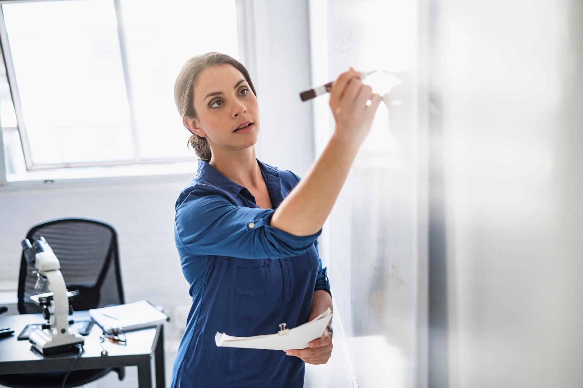 A mid-adult Hispanic female nurse researcher is writing on a whiteboard in her office.