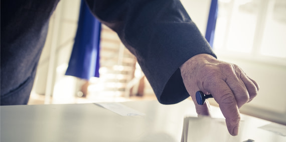 The hand of an older person placing a paper ballot into a box.