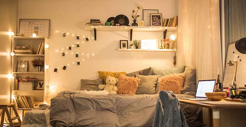 One side of a dorm room is presented in tasteful neutral neutral colors: a simple modern white desk, a bed covered in pillows and a duvet cover, string lights dangling from shelves.