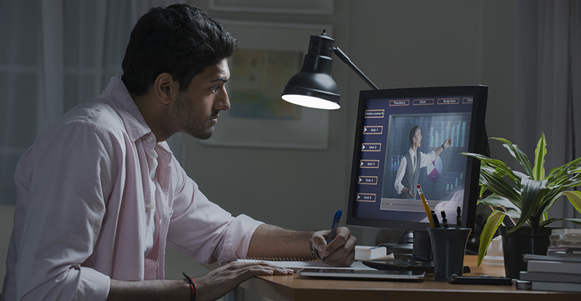 A man in a collared shirt takes notes as he watches a professor lecture in an online class.