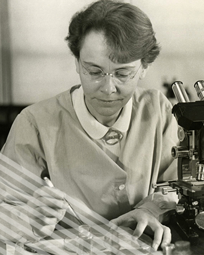 Barbara McClintock, wearing glasses and a lab coat, carefully conducts tests uwith a microscope and petri dishes.