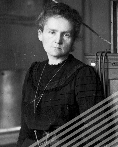 A black-and-white photograph shows Marie Curie in a black dress standing alone in her studio, eyes a little tired, as she looks directly into the camera.