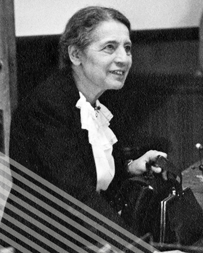Lisa Meitner stands at a table, hand on her purse, as she leans in with a half smile toward someone just off camera.