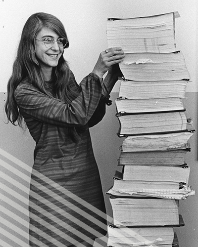 Maragret Hamilton stands next to a stack of code she wrote by hand for the Apollo Project that's almost as tall as she is.