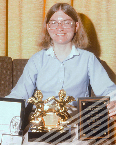 A bespectacled Carol Shaw smiles as she shows off several of awarda for her pioneering video game work.