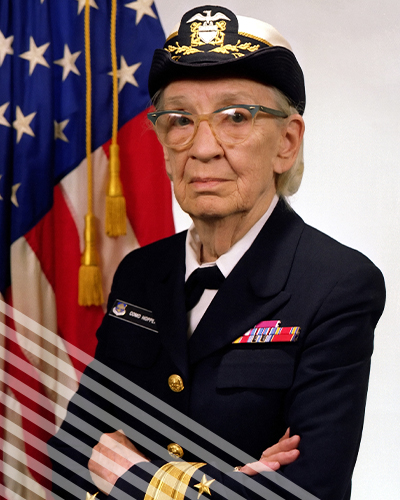 An elderlyGrace Murray Hooper stands in uniform before an American flag.