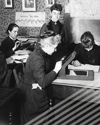 A group of women in full-length dresses pore over calculations in a small room.