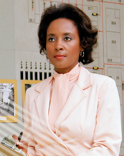 A self-assured Annie Easley in a smart pink blazer takes notes on a clipboard, before a bank of computers.