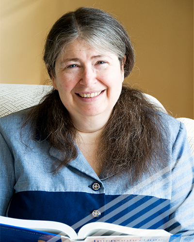 Radi Perlman sits in a livign room with an open book on her lap, smiling at the camera.