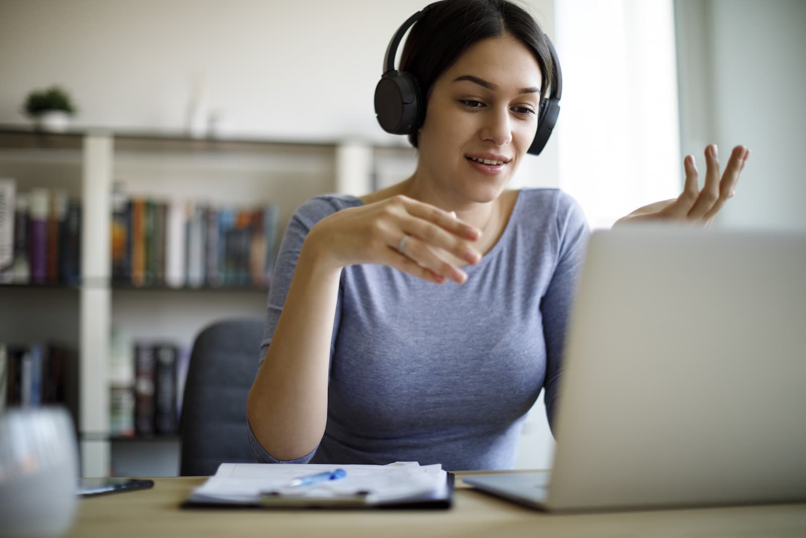 A woman seated at a desk at home wearing headphones gesticulates with her hands as she holds a video call on her laptop computer.