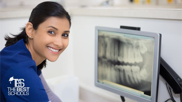 Best Online Dental Assistant Programs (image of woman smiling at camera as she sits in front of a computer displaying an X-ray of a patient’s teeth)