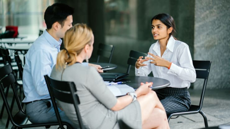 Most Affordable Online MBA Program Hero Image: Most Affordable Online MBA Programs (image of a young woman in business attire animatedly discussing an upcoming business venture with two colleagues seated at an outdoor patio table)