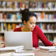Black female college student sitting at a desk in a library on campus. She has her laptop open, and is writing notes in a notebook with a pencil.