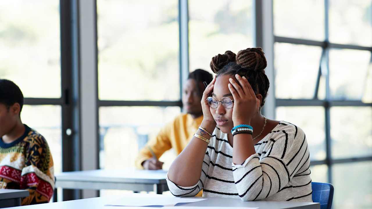 Worried young female student sitting with head in hands at desk during exam in community college classroom