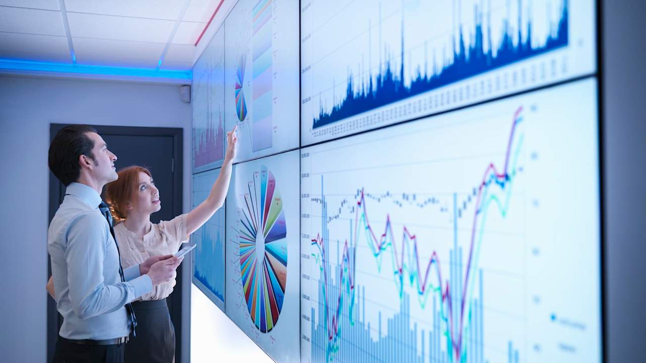 Two colleagues looking at multiple screens showing different types of charts and graphs
