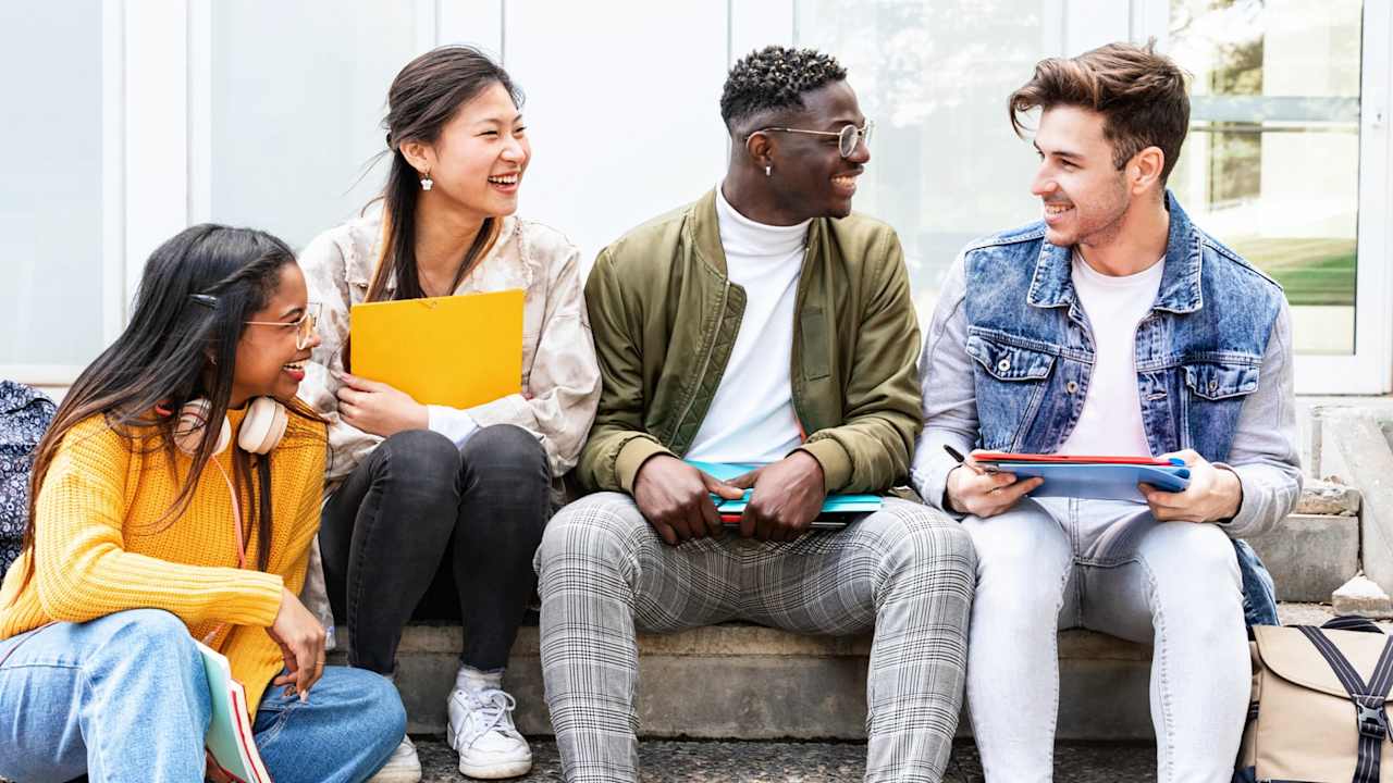 A multicultural group of four friends sitting on steps outside of a university campus building. They are laughing and chatting between classes.