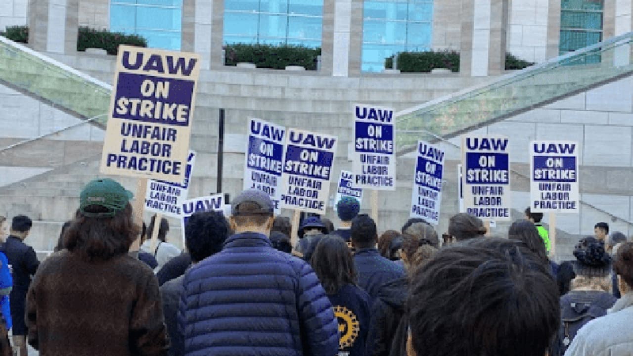 A group of protesters standing out in front of a building with signs that read "UAW on strike, Unfair Labor practices".